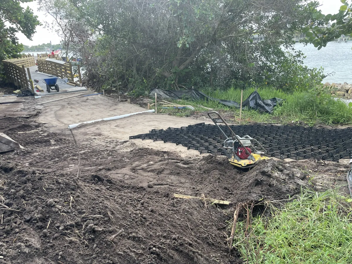 Graded sand and a black plastic ground grid laid for a path, with a plate compactor, leading toward the wooden boardwalk railings at the water's edge at the Jupiter Inlet Lighthouse natural area.